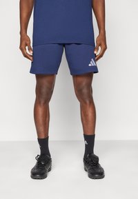 Άνδρας που φοράει σορτσάκι Adidas σε σκούρο μπλε, κάλτσες Adidas μαύρου χρώματος και μαύρα ποδοσφαιρικά παπούτσια, στέκεται μπροστά σε ένα απλό λευκό φόντο.