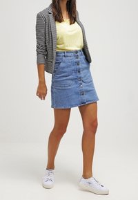 Denim minirok met knopen aan de voorkant, twee opgestikte zakken en een lichtblauwe kleur. Gecombineerd met een gestreepte blazer en een geel topje, inclusief witte sneakers.