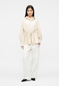 Γυναίκα που φοράει μπεζ πουκάμισο με κουμπιά και μακριά μανίκια, με ζώνη στη μέση, άσπρο παντελόνι με φαρδύ πόδι και μαύρα μυτερά παπούτσια, στέκεται μπροστά σε λευκό φόντο.