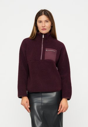 Femme portant un pull polaire bordeaux avec une fermeture éclair à moitié et une poche poitrine zippée, associée à une jupe en cuir noire.