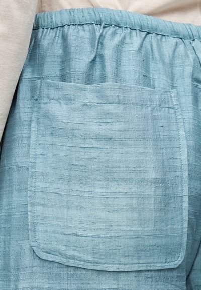 Ανοιχτό μπλε παντελόνι από υφή υφάσματος με ελαστική μέση και μεγάλη τσέπη εμπρός, φορεμένο πάνω από μπεζ ρούχα.