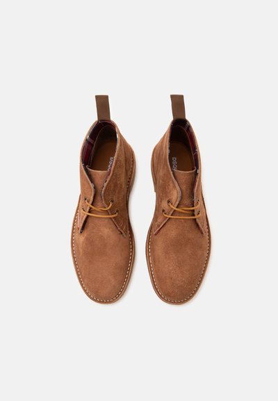 Paire de bottines en daim marron avec lacets en cuir et languettes, vues de dessus sur un fond clair uni.