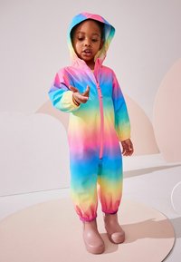 Multicolore regenpak met een verloop van roze naar blauw, voorzien van een capuchon en elastische manchetten. Het pak is gecombineerd met roze rubberen laarzen.
