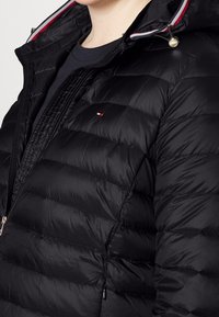 Sort puffjakke med quiltet tekstur, fuld lynlås og snørejusterbar hætte. Har et lille logo samt kontrasterende røde og hvide detaljer.
