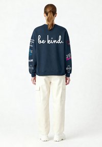 Sērfojuma džemperis tumski zils ar baltu uzrakstu "būt laipnam." aizmugurē, izšūti grafiskie motīvi uz piedurknēm, saskaņots ar krēmkrāsas kravas biksēm un baltām apaviem.