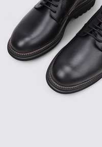 Une paire de chaussures habillées en cuir noir à lacets avec des embouts ronds et des coutures détaillées le long des bords de la semelle sur un fond clair.