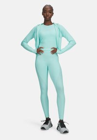 Športna obleka v metežno zeleni barvi, ki vključuje oprijet zgornji del z dolgimi rokavi in visokim pasom na legicah z teksturiranim vzorcem valov, kombinirana s sivimi supergami.