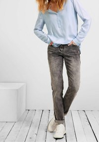 Femme portant une chemise à rayures bleu clair à manches longues, un jean noir délavé et des baskets blanches, posant avec les mains dans les poches sur un sol en bois.