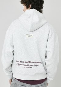 Hoodie gris clair avec capuche à cordon de serrage, poche kangourou et texte brodé en bordeaux foncé dans le bas du dos. Étiquette dans le haut du dos.