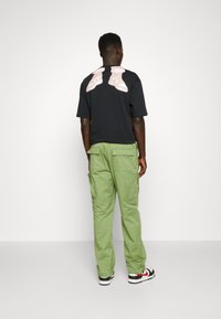 Zwarte T-shirt met een roze sneakerafbeelding op de achterkant, gecombineerd met groene cargobroeken, die een ontspannen pasvorm en meerdere zakken laten zien.