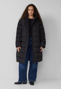 Schwarze Daunenjacke mit Kapuze, im gesteppte Textur, mit Reißverschluss und Seitentaschen, kombiniert mit weit geschnittenen blauen Jeans und schwarzen Stiefeln.