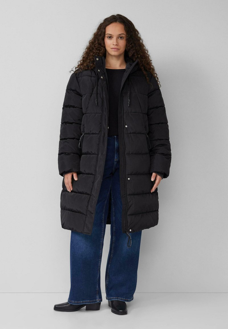 Schwarze Daunenjacke mit Kapuze, im gesteppte Textur, mit Reißverschluss und Seitentaschen, kombiniert mit weit geschnittenen blauen Jeans und schwarzen Stiefeln.