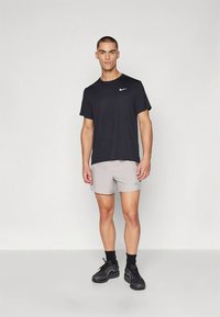 Musta, lyhythihainen Nike T-paita ja vaaleanharmaat shortsit, yhdistettyinä mustiin urheilukenkiin, seisomassa yksinkertaista taustaa vasten.