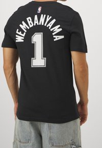 Schwarzes Baumwoll-T-Shirt mit dem Namen "WEMBANYAMA" und der Nummer "1" in großen, stilisierten weißen Buchstaben auf der Rückseite. NBA-Logo am Halsausschnitt.