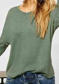 Grön stickad tröja med rund halsringning och avslappnad passform, har ribbad textur och långa ärmar, kombinerad med blå denimjeans.