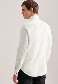 Camicia bianca aderente con maniche lunghe, dotata di colletto strutturato, dettagli con bottoni e design posteriore sagomato. Texture liscia, accenti minimi.