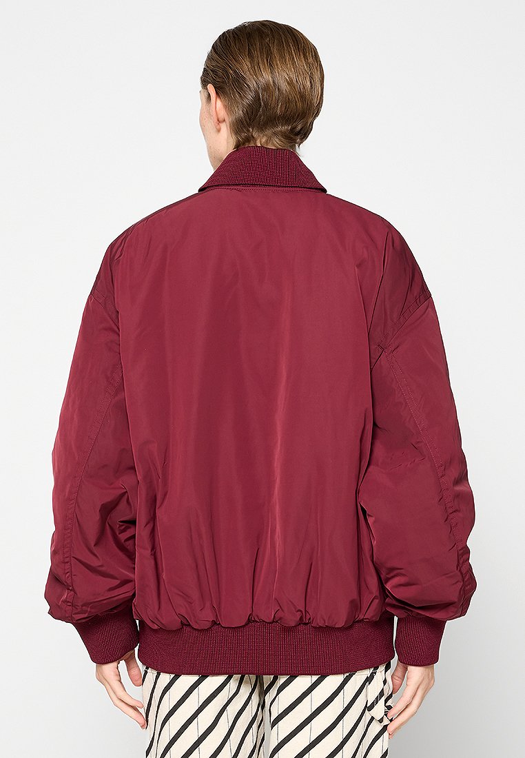 Veste bomber bordeaux avec col et poignets côtelés, offrant un matériau lisse et léger ainsi qu'un design légèrement rembourré. Pantalons rayés visibles.