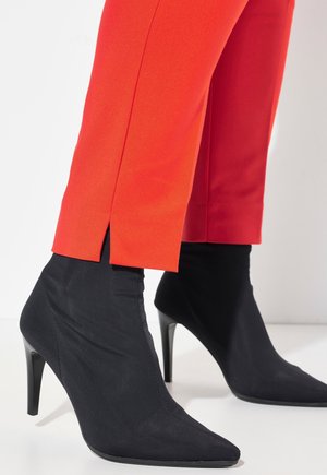 Czarne, spiczaste botki za kostkę o gładkim tekstylnym wykończeniu i szpilką, w połączeniu z jasnoczerwonymi, dopasowanymi spodniami z bocznym rozcięciem.