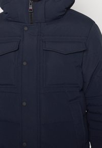 Veste matelassée bleu marine avec capuche, fermeture éclair à l'avant, boutons-pression et deux poches poitrine à rabat avec détails de coutures.