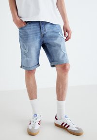 Denim šortky v světle modré barvě s odřenými detaily, ohrnutými manžetami a designem s pěti kapsami, doplněné bílými teniskami a ponožkami do půli lýtek.