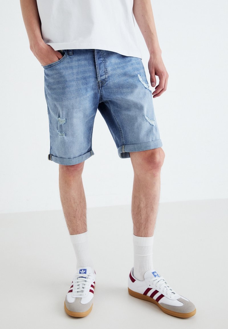 Denim šortky v světle modré barvě s odřenými detaily, ohrnutými manžetami a designem s pěti kapsami, doplněné bílými teniskami a ponožkami do půli lýtek.