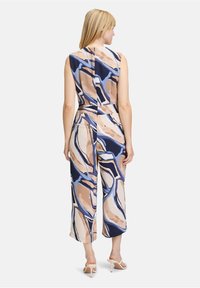 Ujjatlan overall, amely texturált, absztrakt mintával rendelkezik tengerészkék, bézs és kék színekben, egyenes lábcut-tal és hátsó cipzáras részlettel.