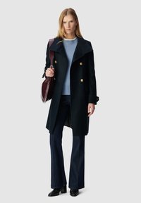 Abrigo cruzado de un oscuro color navy con botones dorados, usado sobre un suéter azul claro y jeans oscuros de campana. Los accesorios incluyen un bolso burdeos.