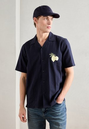 Chemise courte à manches bleu marine en tissu texturé, avec une broderie de ananas jaune sur la poche gauche de la poitrine. Associée à un jean bleu.