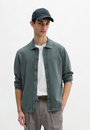 Junger Mann mit dunkelblauer Kappe, offenem türkisfarbenem Hemd über einem weißen T-Shirt, grauer Hose und braunem Gürtel, stehend vor weißem Hintergrund.