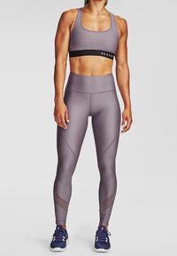 Reggiseno sportivo di colore viola chiaro con fascia nera sottostante; leggings a vita alta dello stesso colore con pannelli testurizzati; abbinati a scarpe da ginnastica blu.