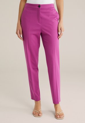 Femme portant un pantalon rose vif ajusté longueur cheville avec bouton et fermeture éclair, associé à des sandales beiges à bout ouvert et un haut blanc.