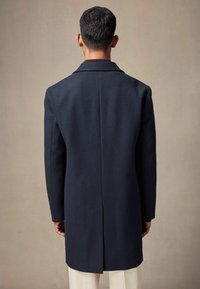 Abrigo de lana azul marino con un corte entallado, que presenta un cuello con muescas, una apertura central y mangas largas, visto desde la parte posterior.