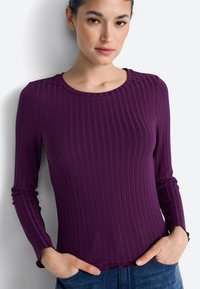 Long-sleeve purple top with a ribbed texture, round neckline, and fitted design, paired with blue jeans. Hands resting on hips.