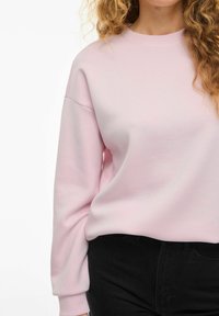 Svetlo roza pulover z okruglim izrezom, sproščenimi rameni in rebrastimi manžetami, v kombinaciji z črnimi hlačami. Gladka tekstura, sproščen dizajn.