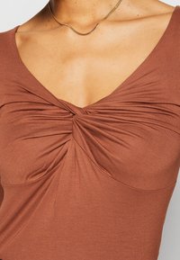 Kobieta w rdzawej bluzce z węzłem na dekolcie w kształcie litery U i cienkim złotym naszyjnikiem.