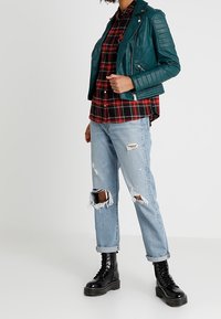 Giacca biker in pelle teal con dettagli trapuntati, camicia a quadri rossa e nera, jeans chiari strappati e stivaletti neri con lacci chunky.