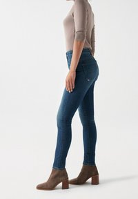 Jeans ajustados de mezclilla azul oscuro con cintura alta, con un acabado texturizado, bolsillos traseros cosidos y combinados con botas de tobillo marrones.