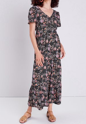 Nainen pukeutuneena mustaan kukkakuosiseen midi-mekkoon, jossa on lyhyet hihat, ja ruskeat sandaalit, seisomassa yksinkertaisen vaalean taustan edessä.