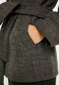 Grå herringbone-mönstrad kappa med bälte, med en sidoficka och en mjuk tygtextur. Accenter ingår i designen.