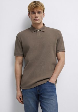 Junger Mann mit blonden Haaren steht vor einer weißen Wand, trägt ein braunes kurzärmeliges Poloshirt und blaue Jeans, eine Hand in der Tasche.