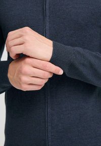 Sötét szürke kötött kardigán cipzárral, bordás mandzsettákkal és sima felülettel. A kezek a ujjak állításán dolgoznak.