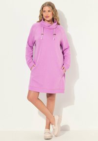 Vestido sudadera rosa de cuello con capucha y mangas largas, con bolsillos delanteros y detalles de cordón. Tejido suave con un corte relajado, combinado con zapatos ligeros.