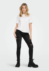 Maglietta bianca a maniche corte abbinata a jeans skinny neri strappati e stivali neri con lacci. L'outfit presenta uno stile urbano e casual.