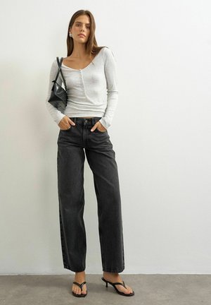 Young woman stands against white wall wearing light grey long sleeve top, black wide-leg jeans, black heeled sandals, and small black shoulder bag.