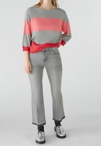 Personne portant un pull rayé gris et corail, un jean gris court avec des ourlets effilochés, des chaussettes noires et des mocassins argentés, se tenant contre un fond blanc.