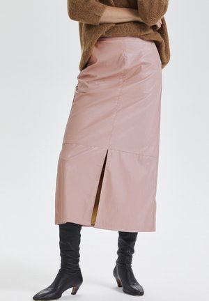 Mujer con falda midi rosa con una abertura en la parte trasera, combinada con botas negras altas con tacón y un suéter marrón.