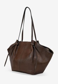 Bolso tote de cuero marrón con una textura suave, forma redondeada y lazos laterales ajustables. Presenta asas largas y planas para un fácil transporte.