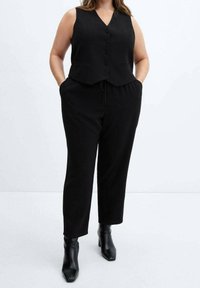 Sort, ærmeløs vest med v-hals og knapper, parret med sorte tapered bukser og ankelstøvler, der skaber et monokromatisk look.