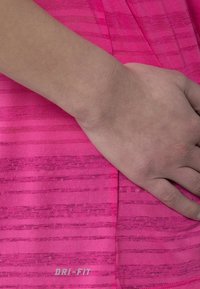 Růžová Dri-Fit tkanina s jemným pruhovaným vzorem, hladkou texturou a viditelným logem na dolní části, s rukou odpočívající na tkanině.