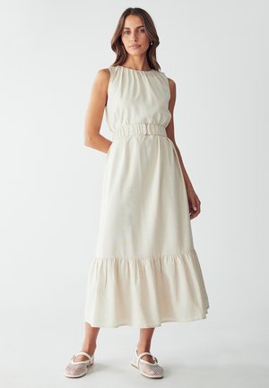 Helles beiges ärmelloses Kleid mit gerafftem Oberteil, elastischem Bund und mehrlagigem Rüschensaum. Kombiniert mit weißen, durchlöcherten flachen Schuhen.